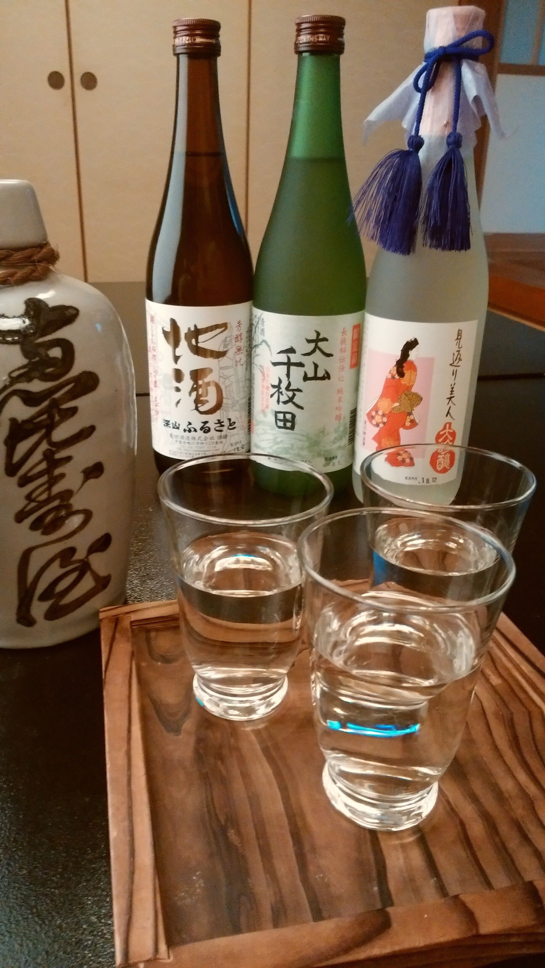 Four bottles of Japanese sake and three shot glasses on a wooden tray.
