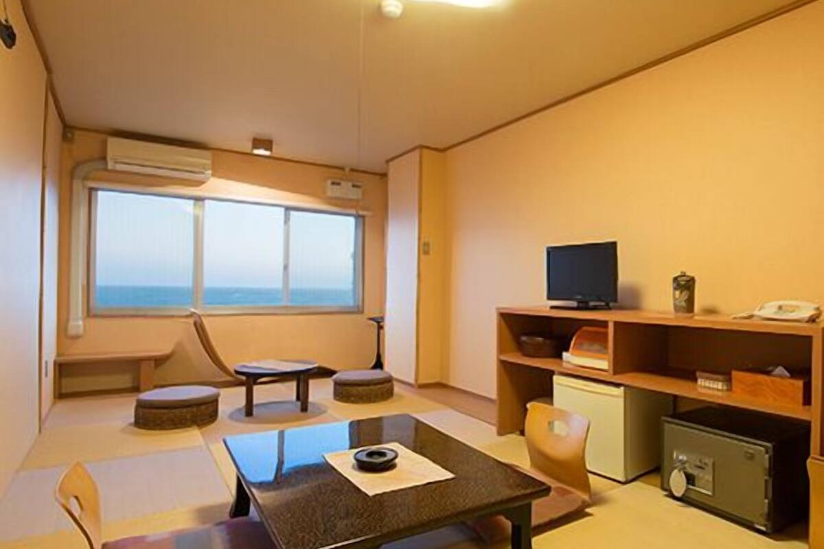 Ein Zimmer mit Tatami-Böden, einem niedrigen Tisch, einer Couch, einem Fernseher und einem kleinen Schrank.