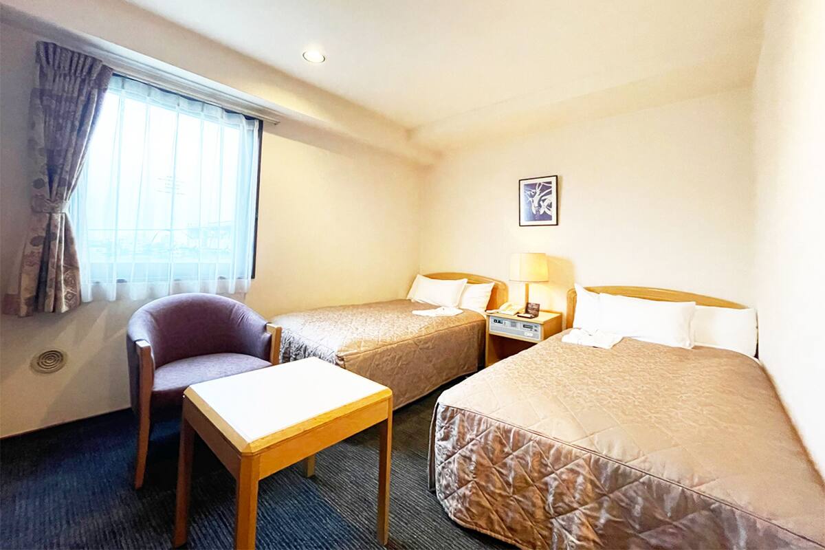 A hotel room with two single beds, a small table, a chair, a desk, and a window with curtains.