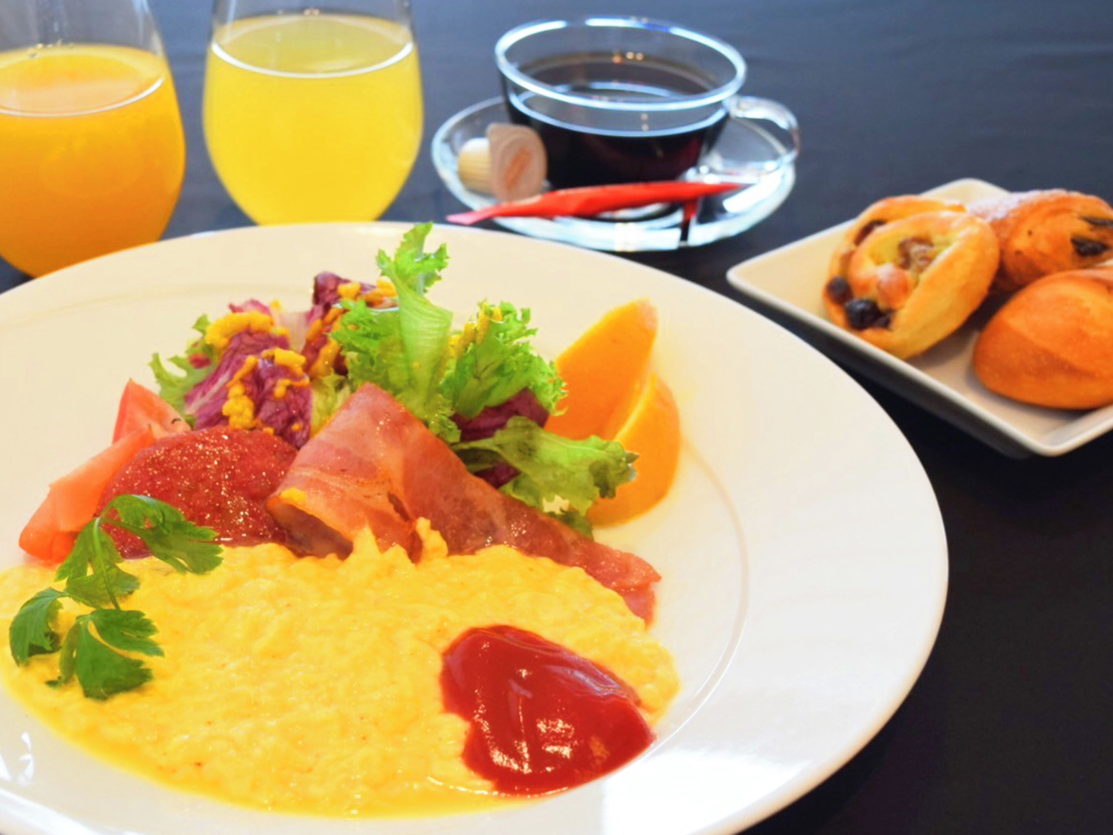 Un plato de desayuno con huevos revueltos, tocino, tomates y una ensalada, acompañado de jugo de naranja y una taza de café.