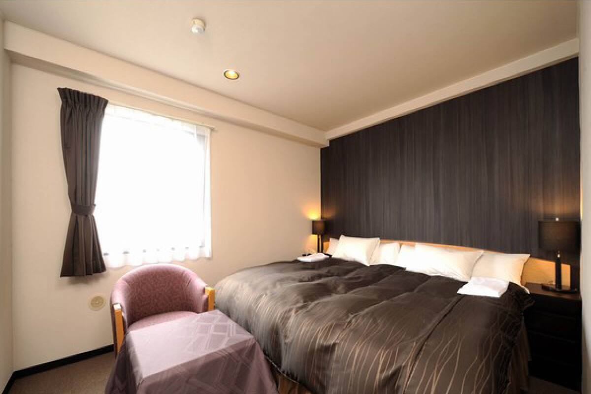 A hotel room with a large bed, a bedside table with a lamp, a chair with a table, and a window with curtains.