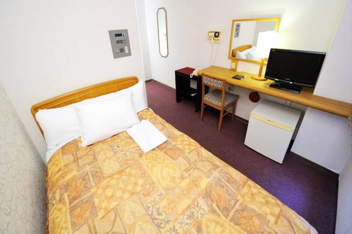 A hotel room with a bed, a desk with a computer, a chair, a small refrigerator, and a mirror.