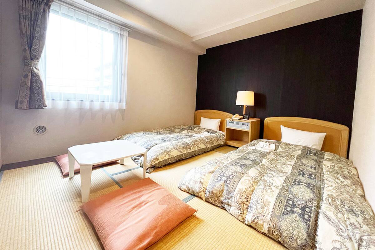 A hotel room with two beds, a small table, a nightstand with a lamp, and a window with curtains.