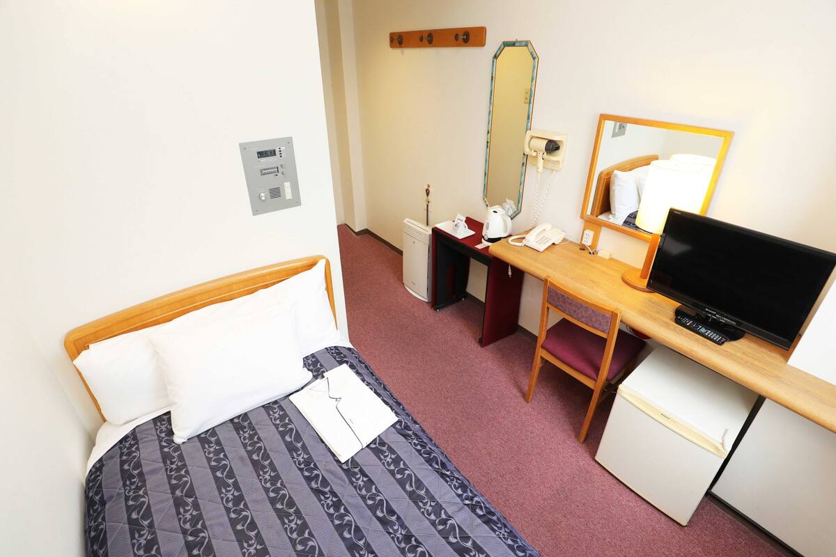 A hotel room with a bed, a desk with a computer, a mirror, and a small refrigerator.