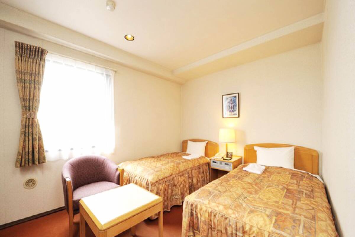 A hotel room with two beds, a small table, a chair, a nightstand with a lamp, and a window with curtains.