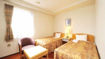 Quarto de hotel com duas camas, uma mesa pequena, uma cadeira, uma mesinha de cabeceira com abajur e uma janela com cortinas.
