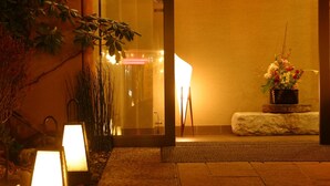 Una entrada bien iluminada con puerta de vidrio, un banco de piedra y una planta con flores.