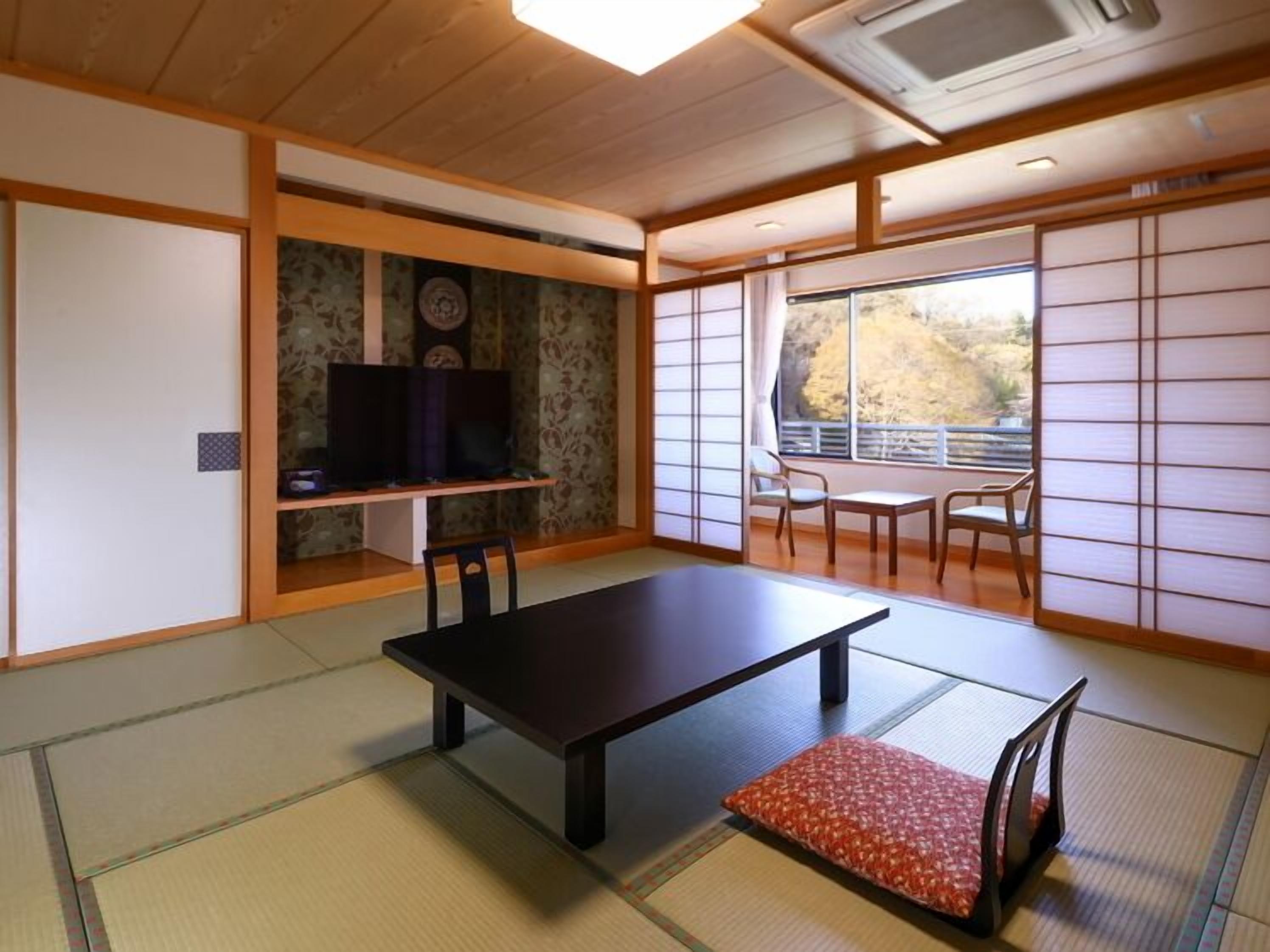 Una habitación japonesa tradicional con suelo de tatami, una mesa baja, sillas, un televisor y puertas correderas.