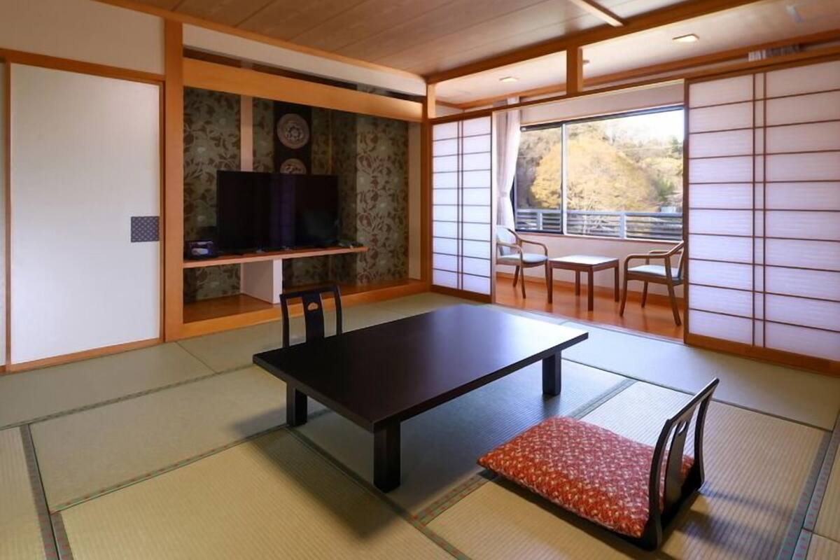 Una habitació japonesa tradicional amb fons de tatami, una taula baixa, cadires, un televisor i portes corredisses.