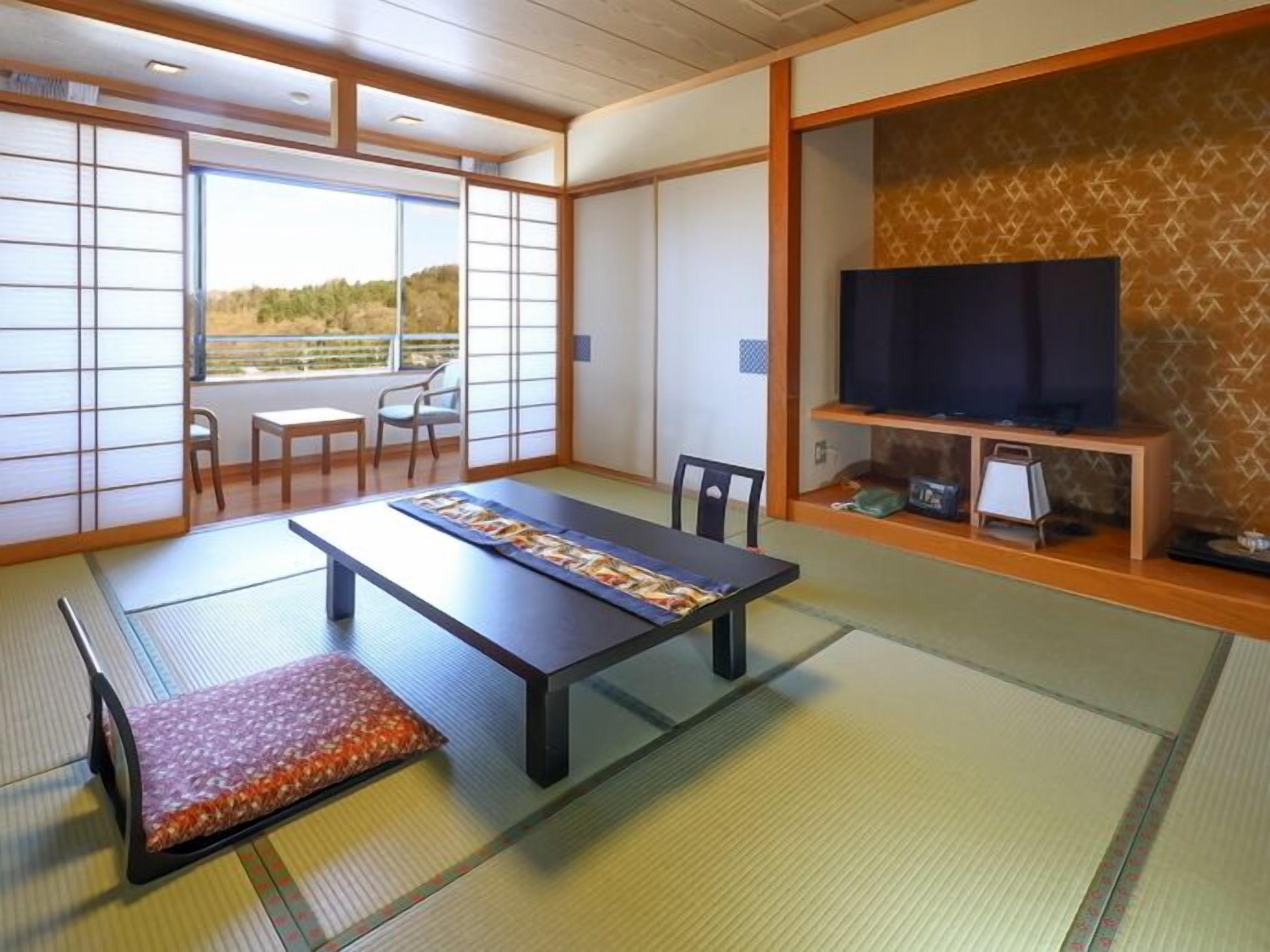 Una habitación con suelo de tatami, una mesa de tatami, un televisor sobre un soporte de madera y vistas a través de un celosía japonés.