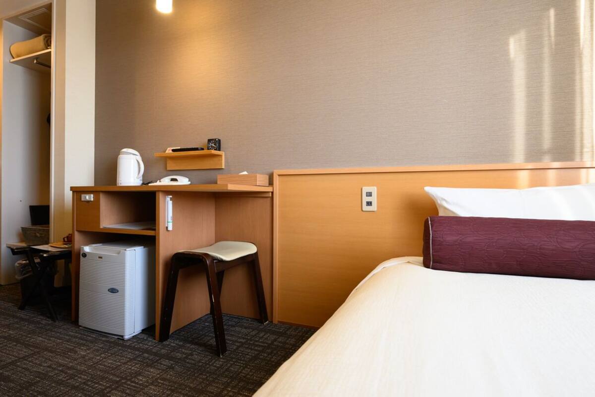 A hotel room with a bed, a desk, a chair, a small refrigerator, a shelf with a kettle, and a bedside table.