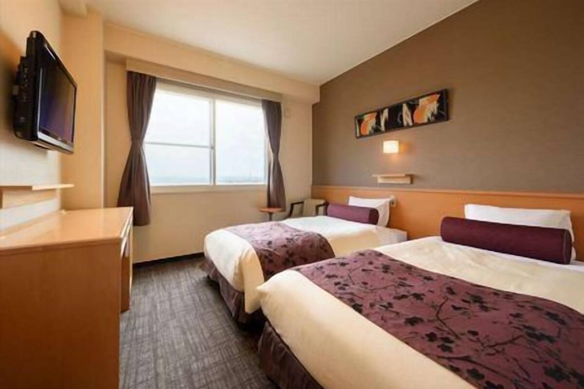 A hotel room with two beds, a desk, a flat-screen TV, a window with curtains, and a painting on the wall.