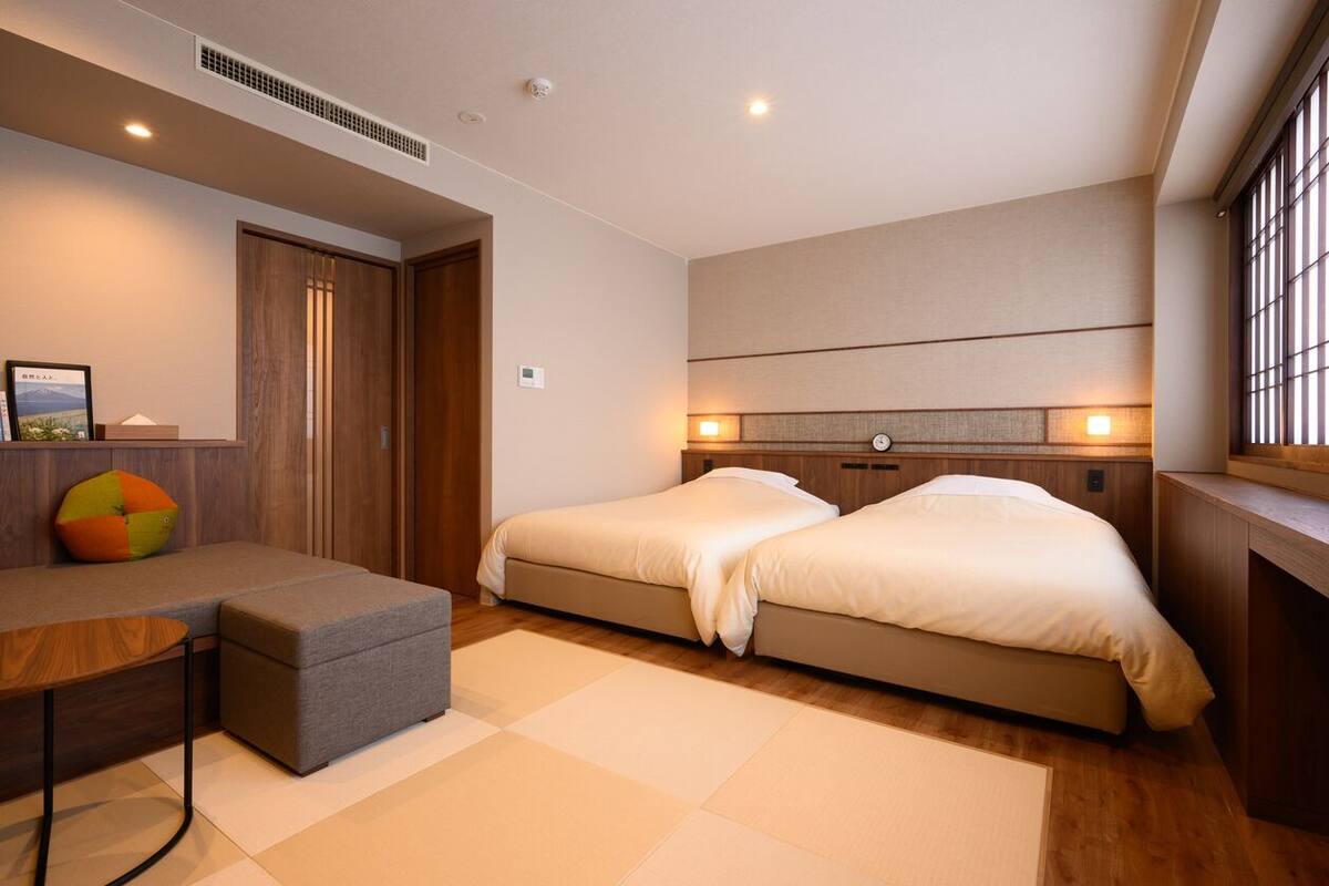 A modern hotel room with a sofa, two beds, a small table, and a window with blinds.