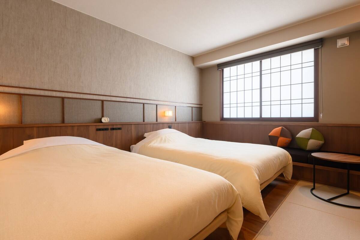 A hotel room with two beds, a window with blinds, a bedside table, and a wall clock.