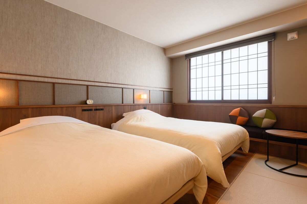 A hotel room with two beds, a window with blinds, a bedside table, and a wall clock.