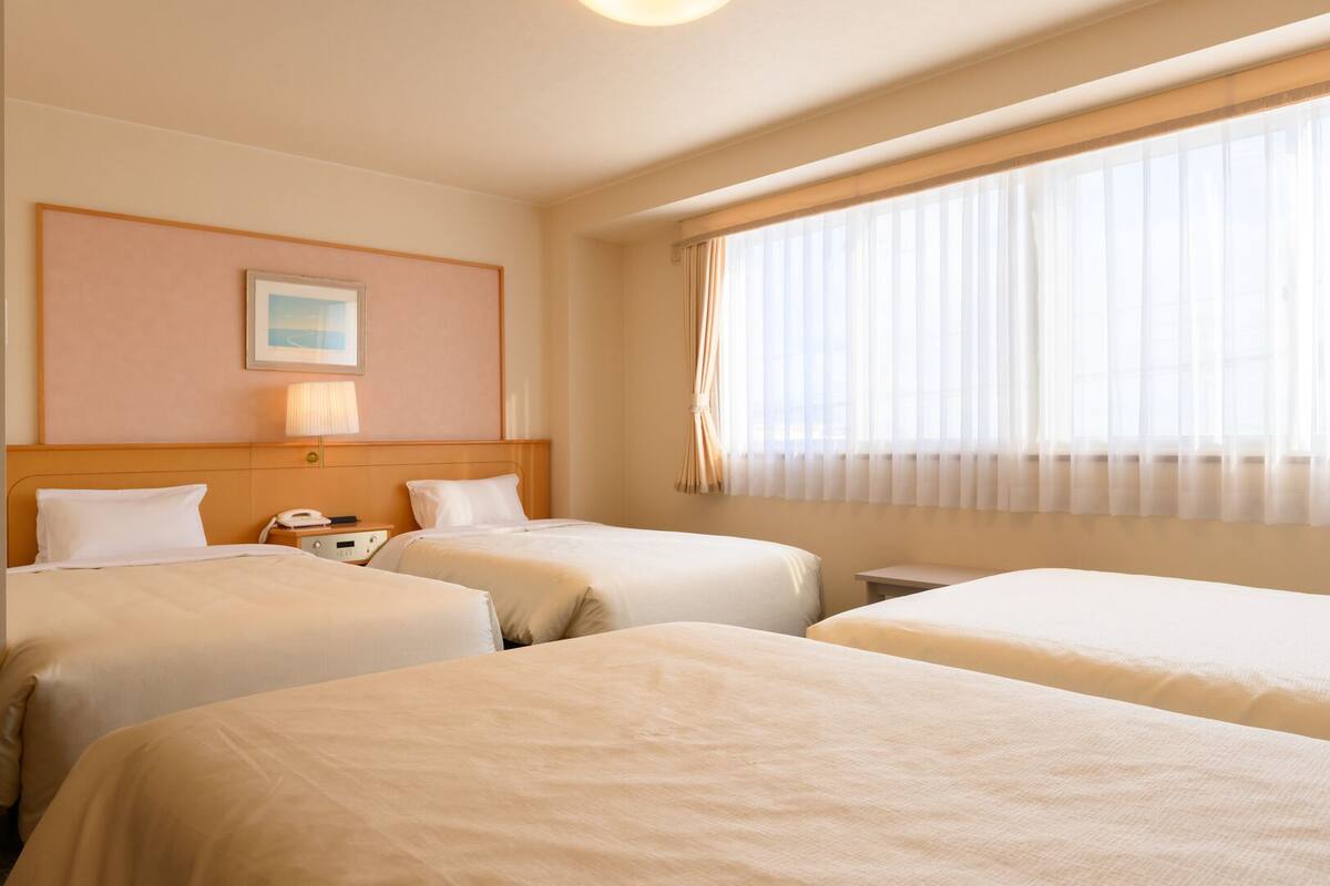 A hotel room with three beds, a bedside table with a lamp, a painting on the wall, and a window with sheer curtains.