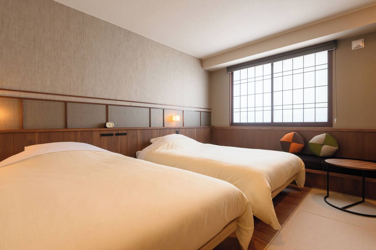 A hotel room with two beds, a window with blinds, a small table, and a wall-mounted heater.
