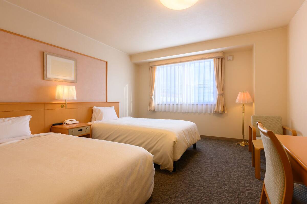 A hotel room with two beds, a desk, a chair, a lamp, a telephone, and a window with curtains.