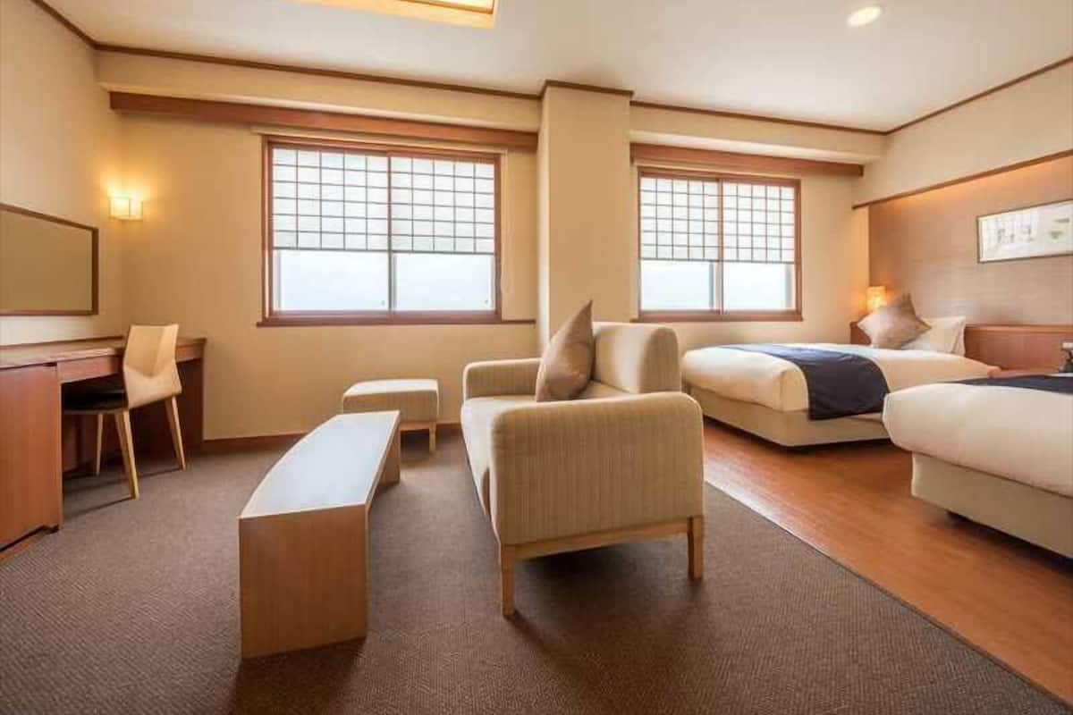 A hotel room with a desk, two couches, a bench, and two windows with blinds.