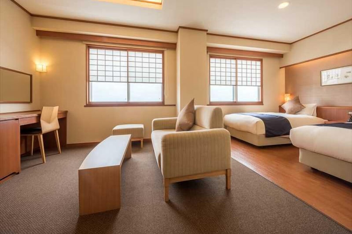 A hotel room with a desk, two couches, a bench, and two windows with blinds.