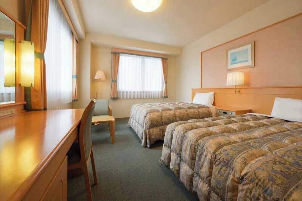 A hotel room with two beds, a desk, a chair, a lamp, a window with curtains, and a painting on the wall.
