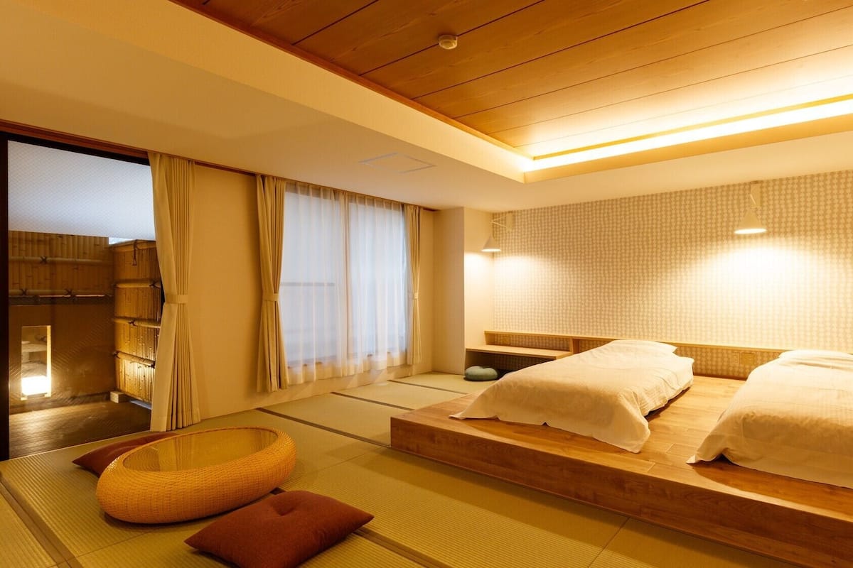 Une pièce de style japonais traditionnel, avec un sol recouvert de tatamis, un lit bas en bois avec une literie blanche, un futon, un panier tressé et une porte coulissante donnant accès à la salle de bain.