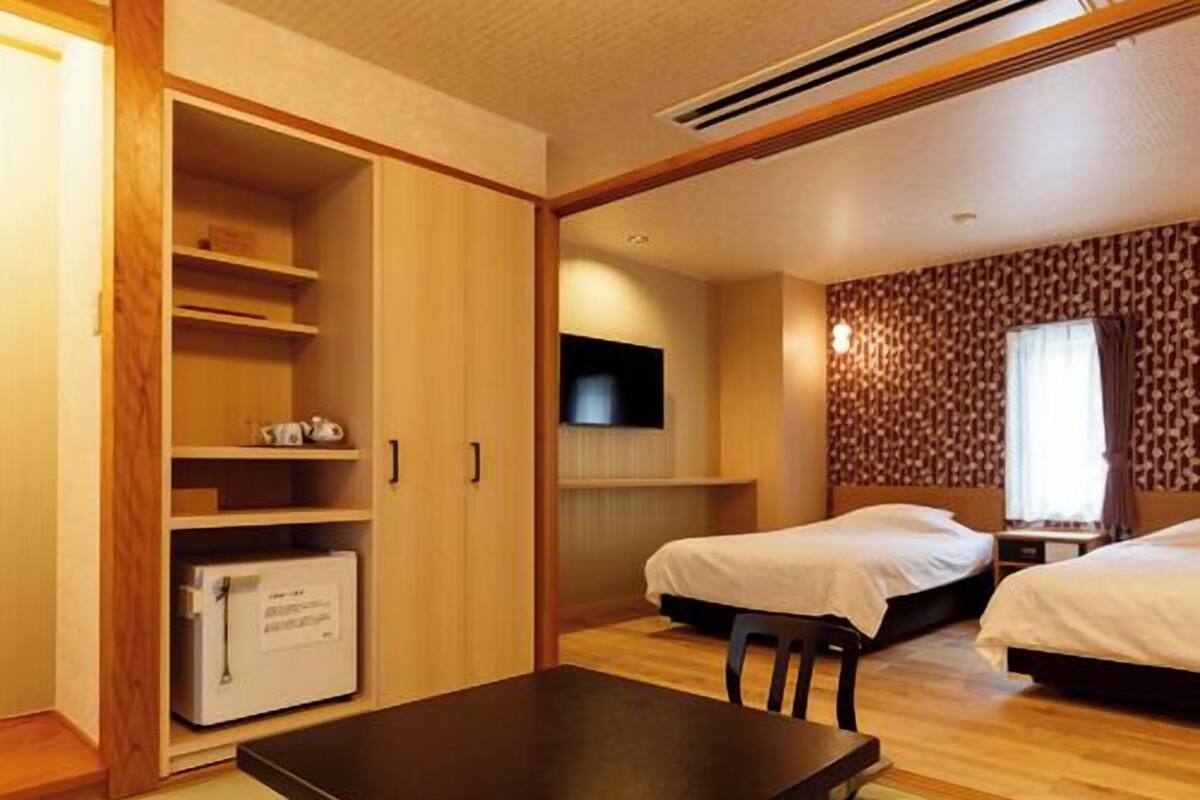 A hotel room with a wooden wardrobe, a desk with a chair, two beds, a flat-screen TV, and a patterned wall.