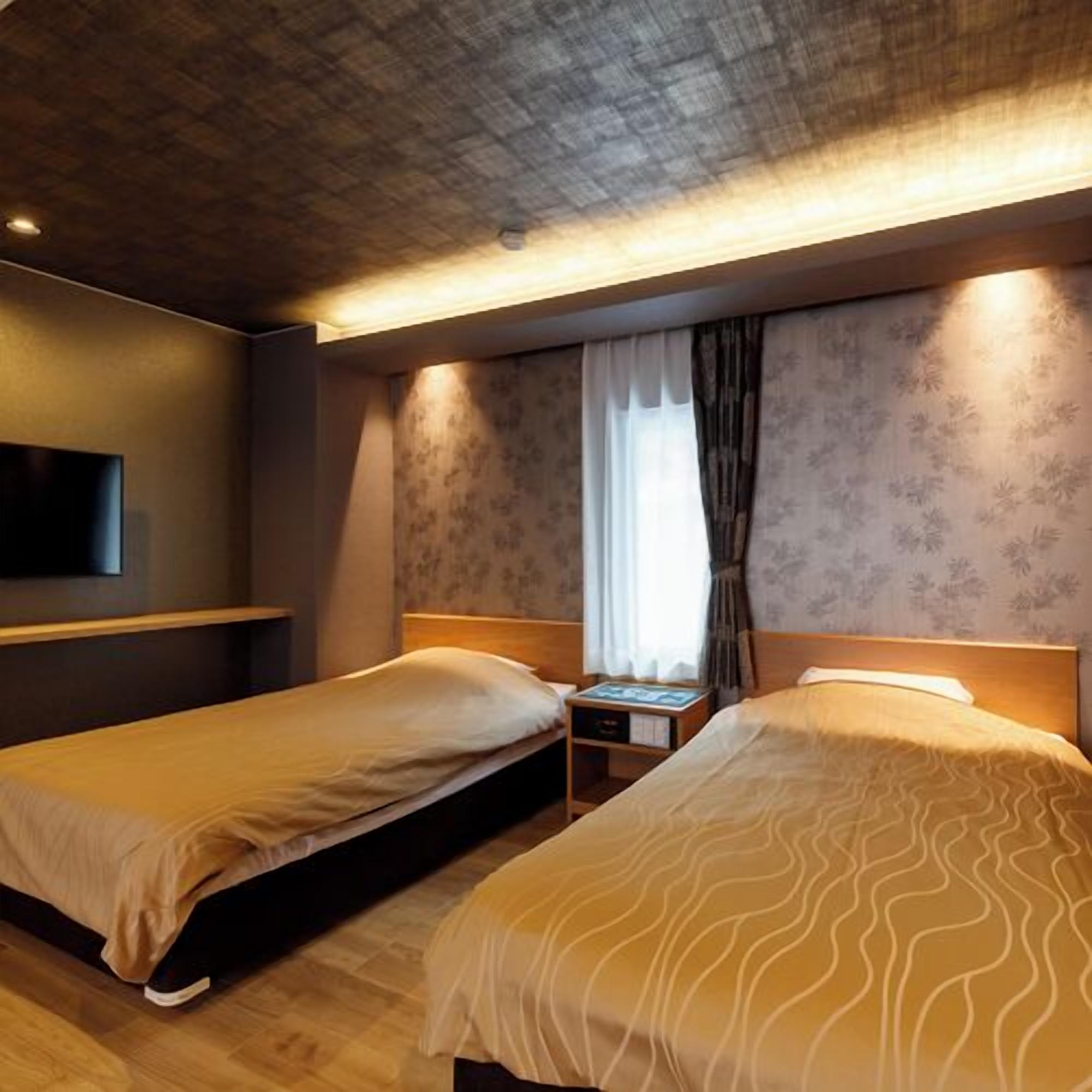 A hotel room with two beds, a TV mounted on the wall, a nightstand, and a window with curtains.