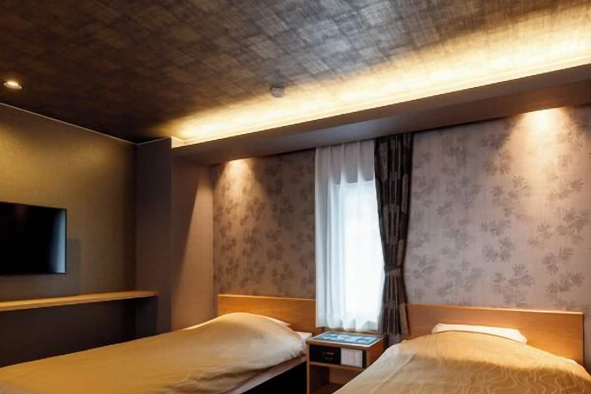 Une chambre d’hôtel avec deux lits, un téléviseur fixé au mur, une table de chevet et une fenêtre avec des rideaux.