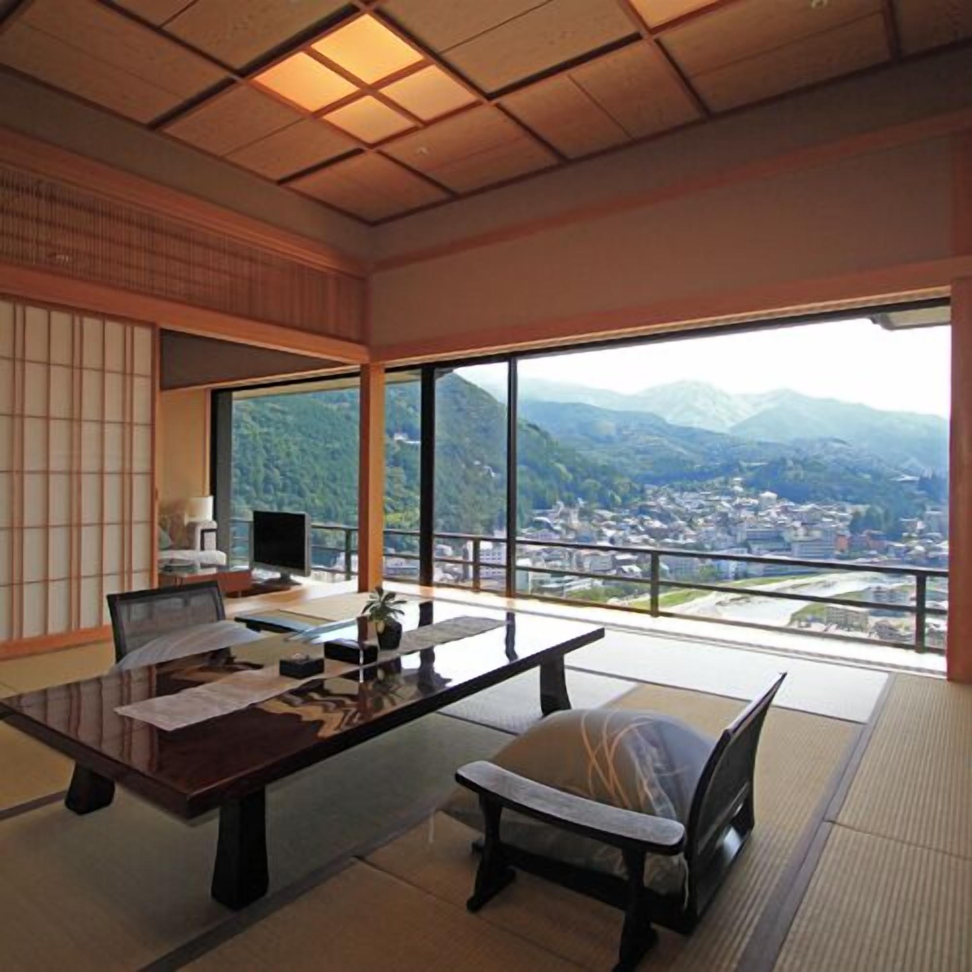 Ein Zimmer mit Tatami-Boden, einem niedrigen Tisch mit Stuhl, einem Schreibtisch mit Stuhl, einem Fernseher und einem großen Fenster mit Blick auf Berge und eine Stadt.