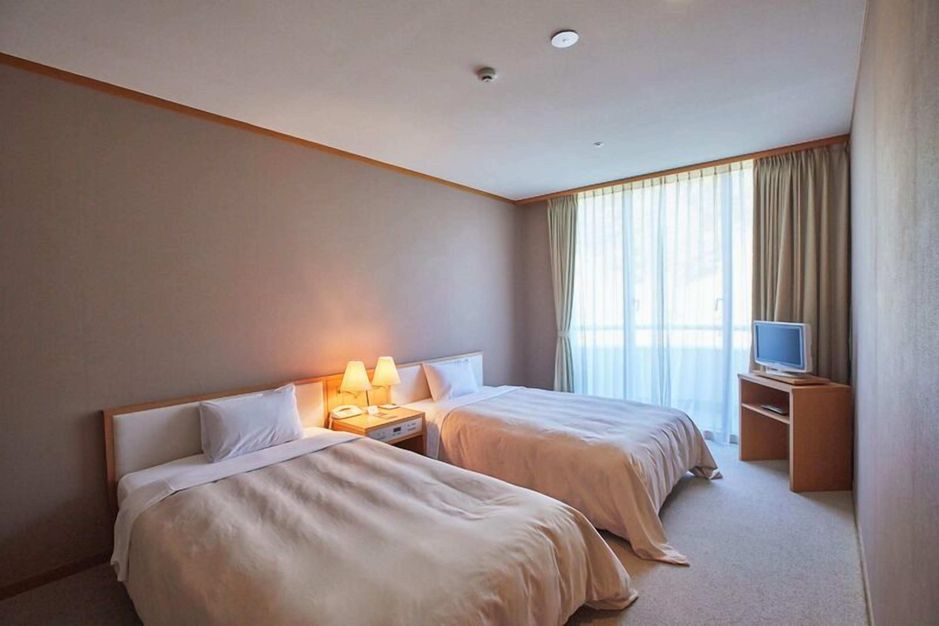 Quarto de hotel com duas camas, televisão, secretária e janela com cortinas.