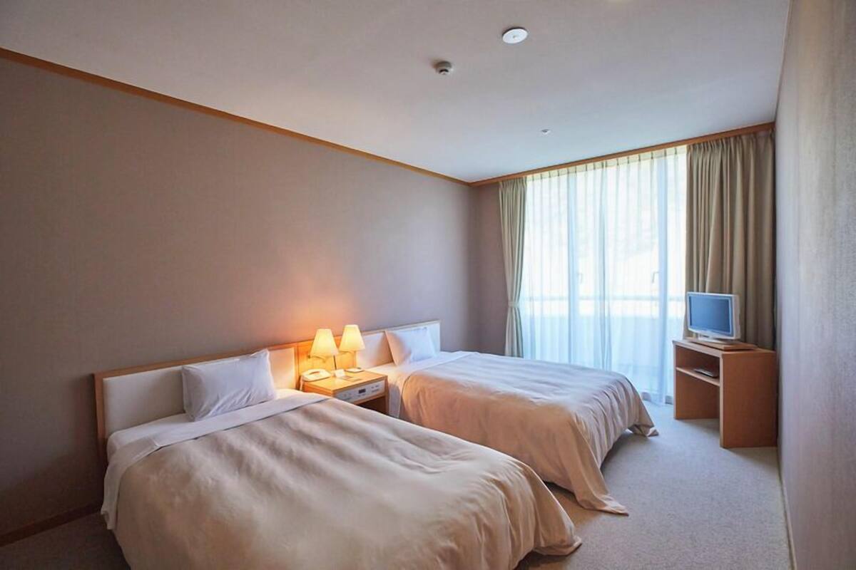 A hotel room with two beds, a TV, a desk, and a window with curtains.
