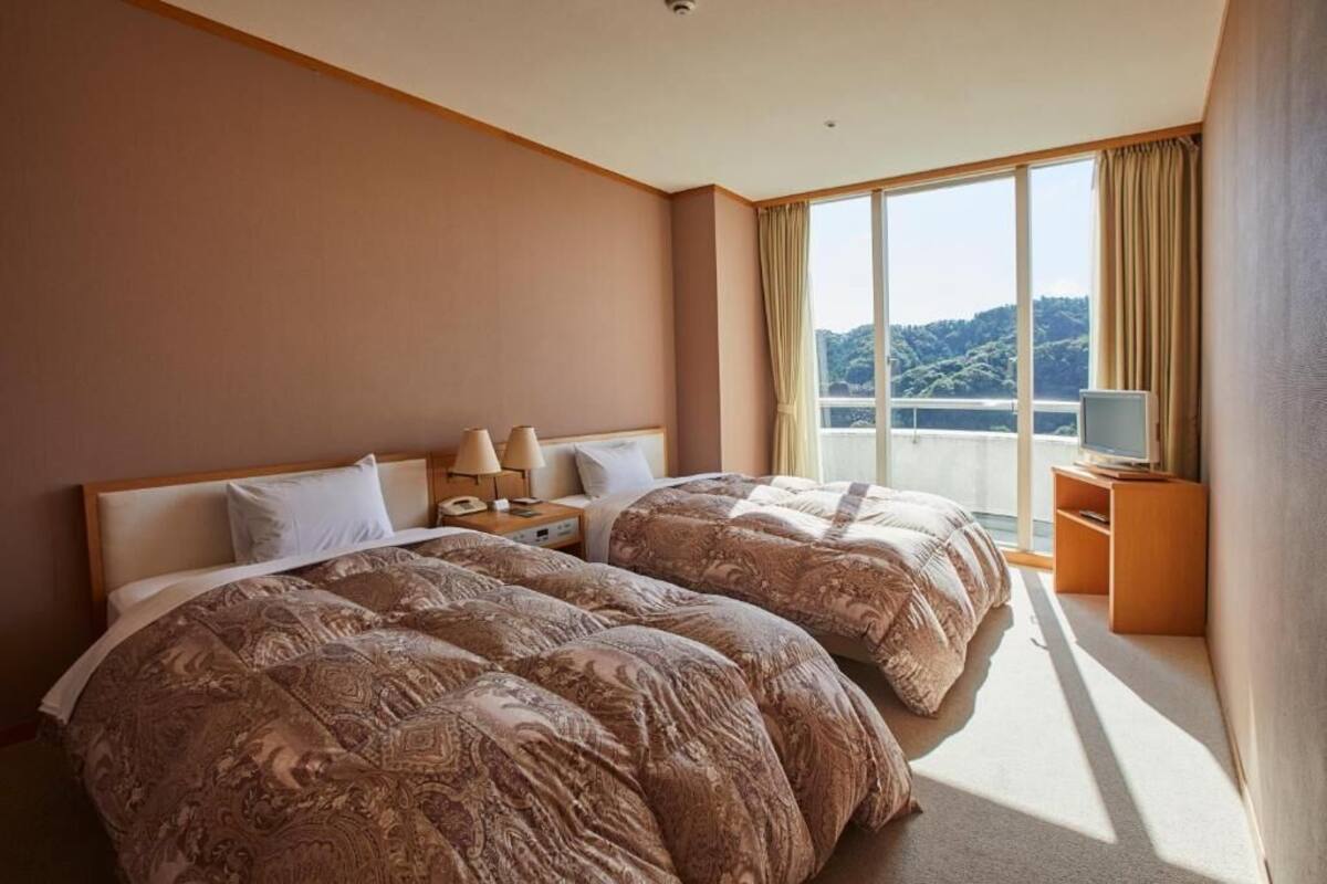 A hotel room with two beds, a large window with a view, a bedside table with a lamp, and a TV on a stand.