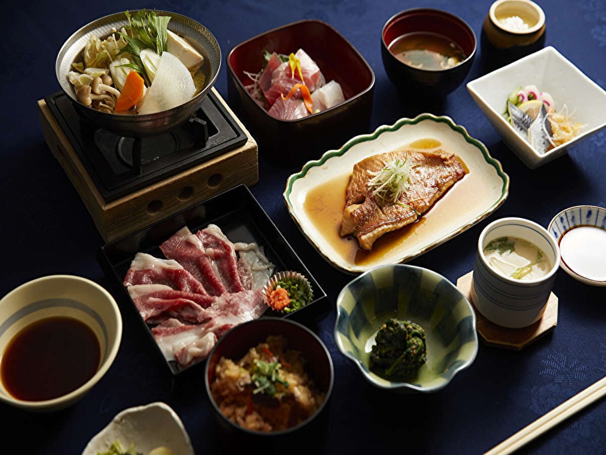A variety of Japanese dishes including sashimi, tofu, and miso soup, arranged on a dark table with chopsticks and small bowls.