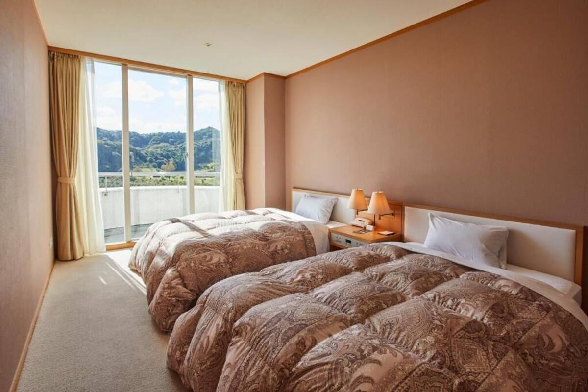 A hotel room with two beds, a large window with curtains, and a view of a mountainous landscape.
