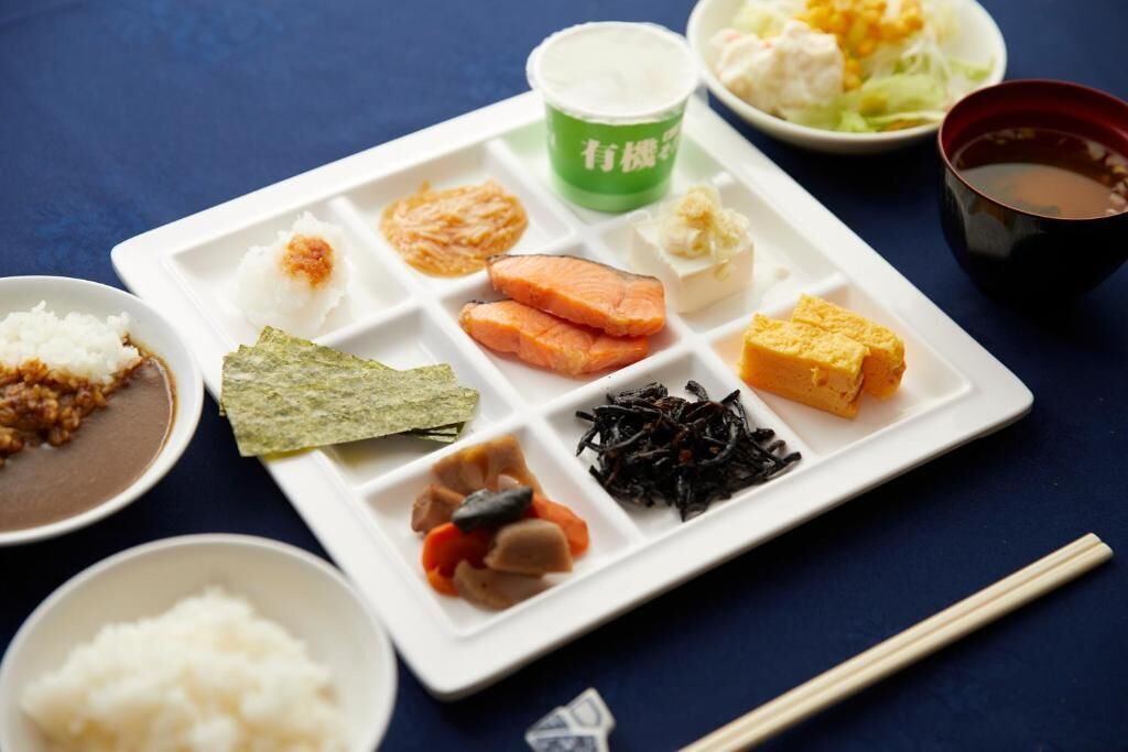 A variety of Japanese dishes on a white plate, including rice, salmon, tofu, and seaweed, with chopsticks and a cup of tea on the side.