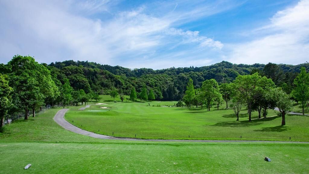 Un campo de golf con un green cuidado, un sendero serpenteante y una colina boscosa al fondo.