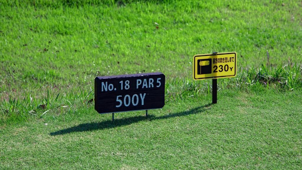 Two signs in a grassy area, one black with white text reading "No. 18 PAR 5 500Y" and another yellow with black text "前方の駐車場が開いて230Y".