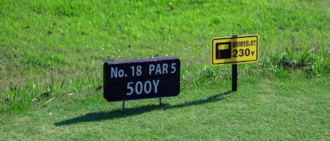 Two signs in a grassy area, one black with white text reading "No. 18 PAR 5 500Y" and another yellow with black text "前方の駐車場が開いて230Y".