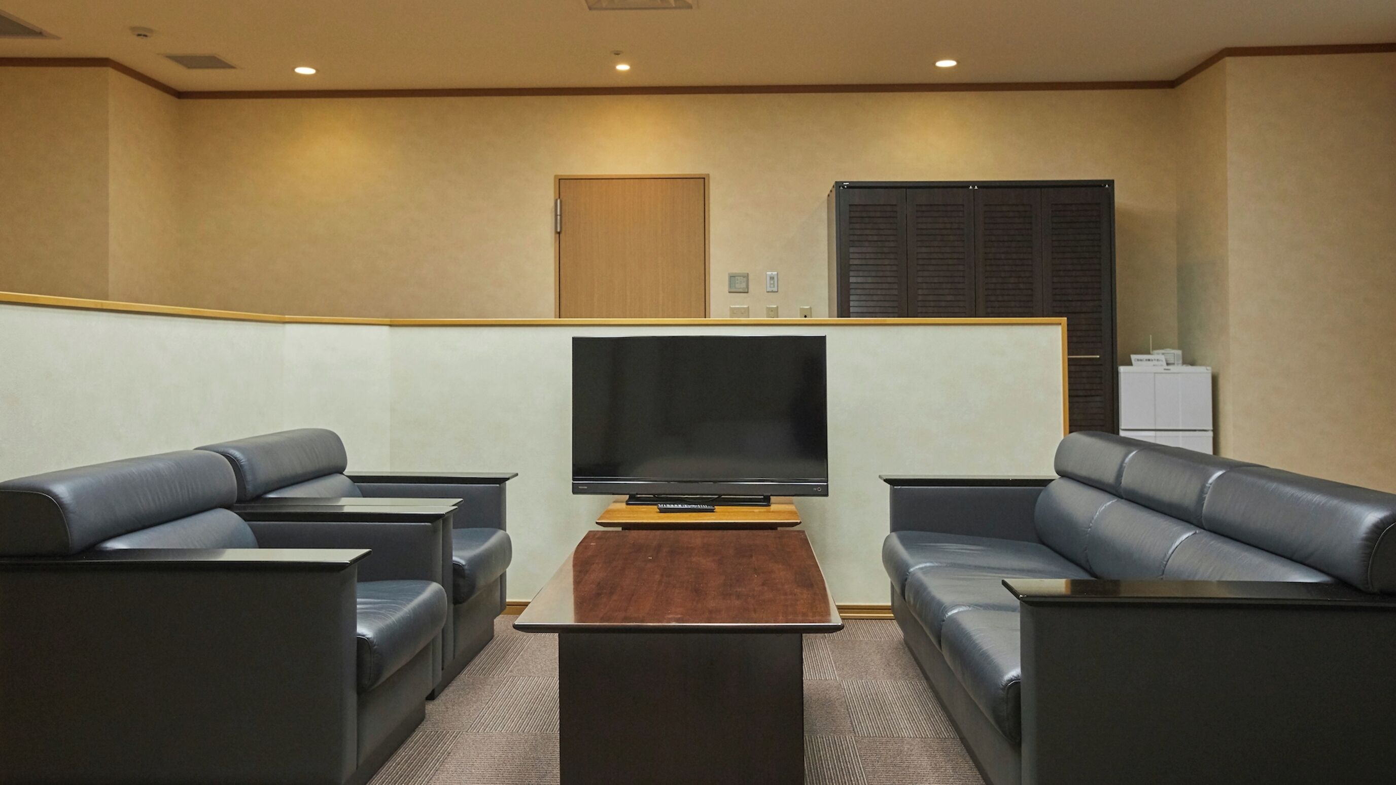 A modern living room with a flat-screen TV, leather sofas, a wooden coffee table, and a built-in cabinet with a refrigerator.