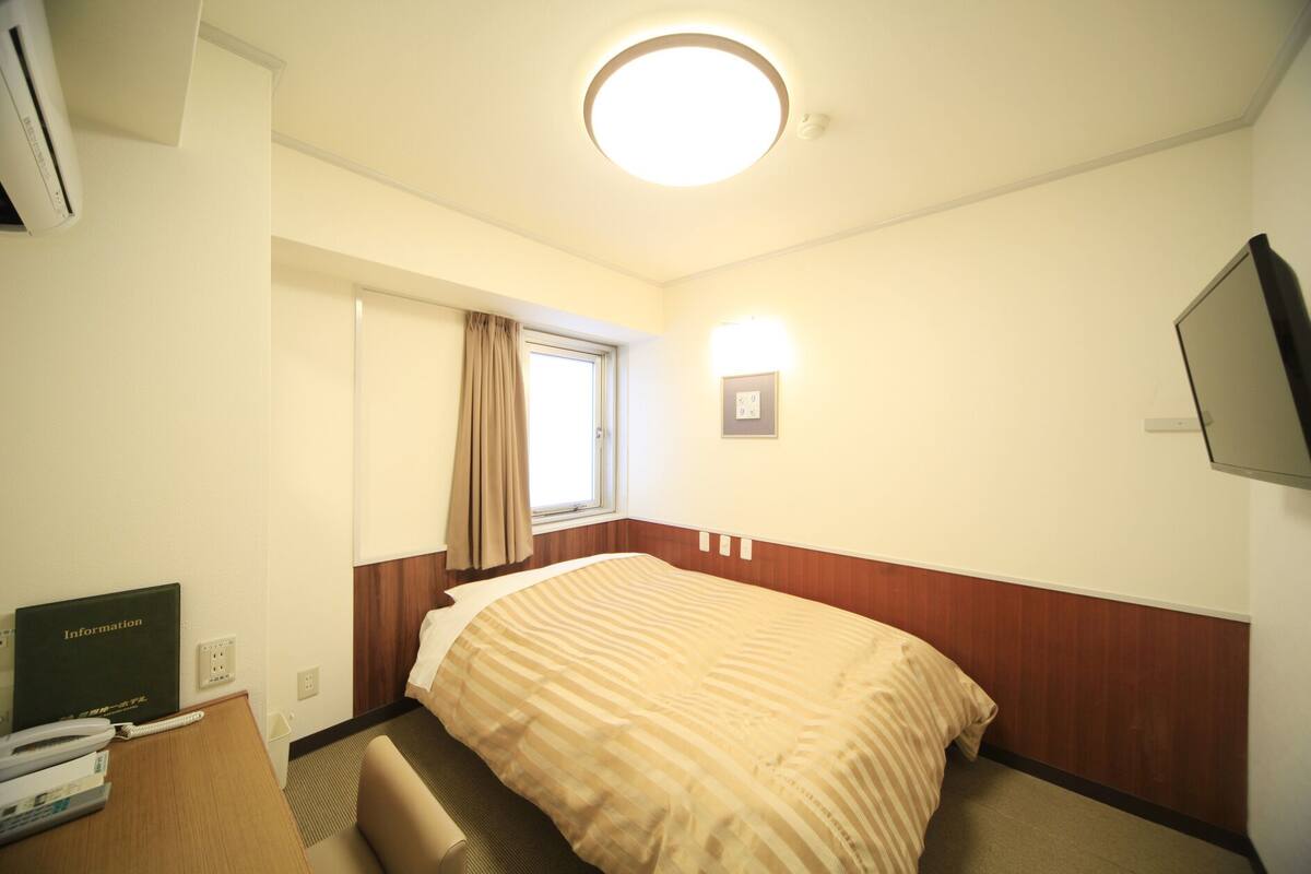 Una habitació d'hotel amb un llit, una taula de nit amb telèfon, televisió i finestra amb cortines.
