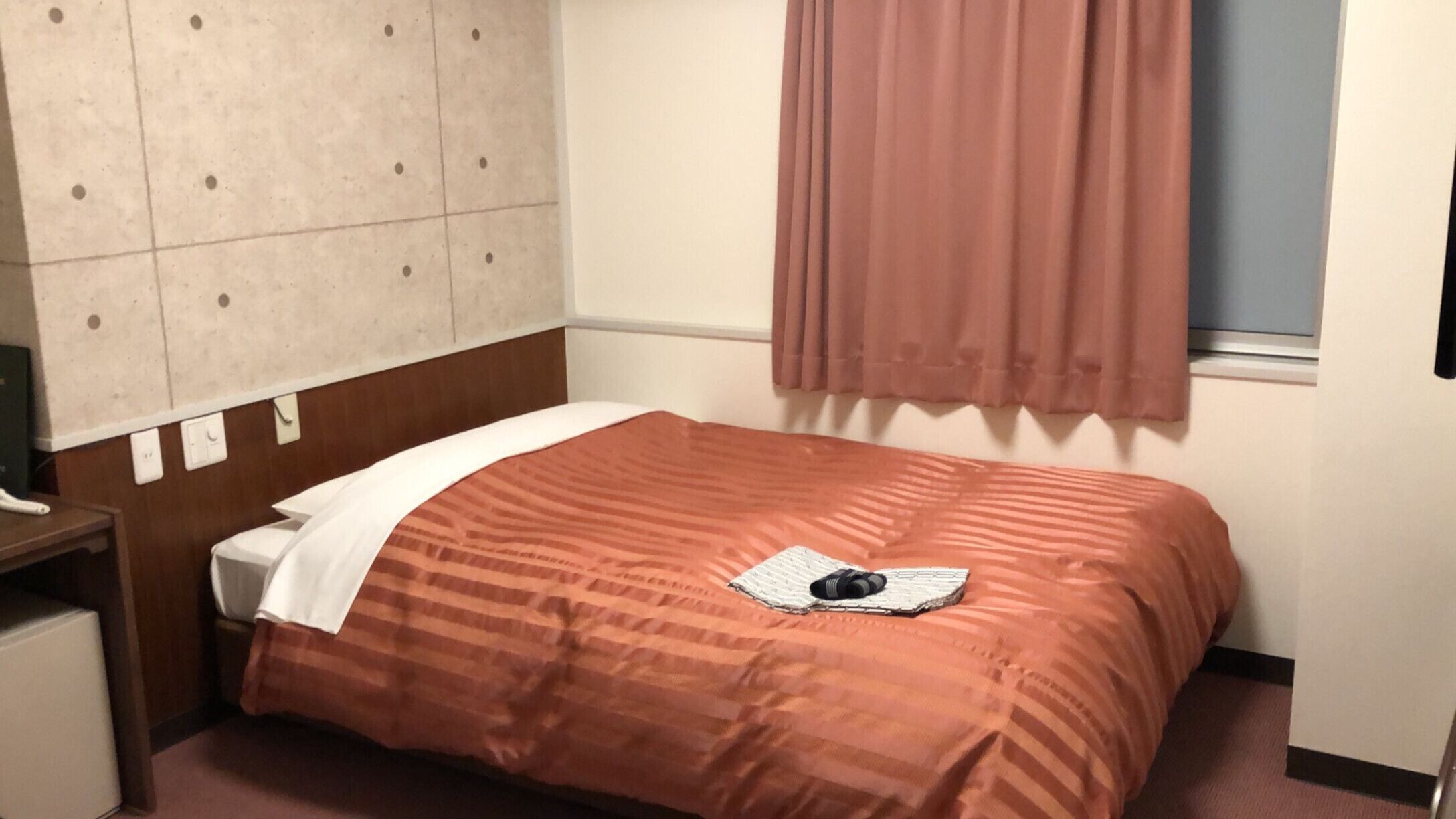 Een hotelkamer met een bed, nachtkastje, raam met gordijnen en een wand met een patroon.