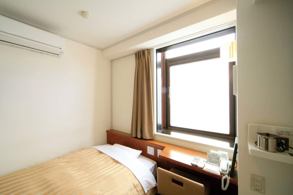 Habitación de hotel con cama, escritorio con teléfono, ventana con cortinas, unidad de aire acondicionado y televisor montado en la pared.