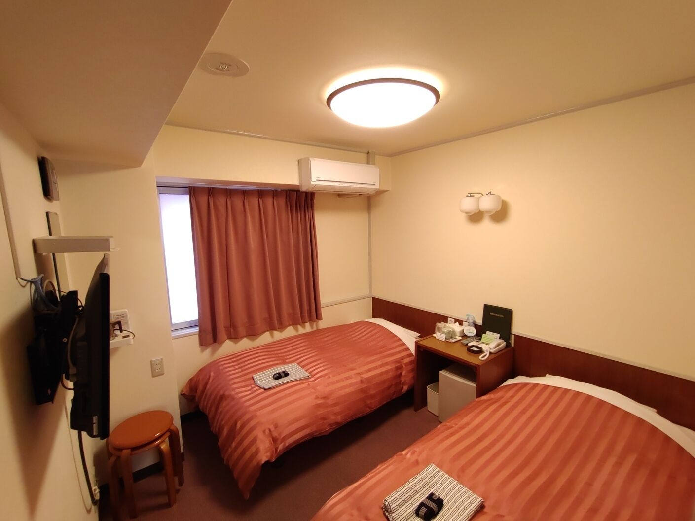Hotelkamer met twee bedden, een bureau, een stoel, een raam met gordijnen, een airconditioning en een wandlamp.
