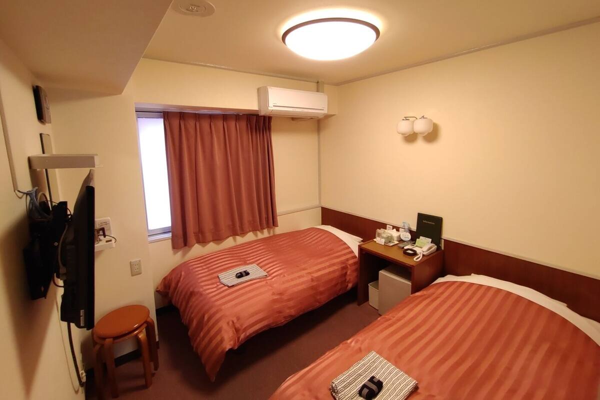 Habitació d'hotel amb dos llits, una taula de treball, una cadira, una finestra amb cortines, un aparell d'aire condicionat i una làmpada penjada a la paret.