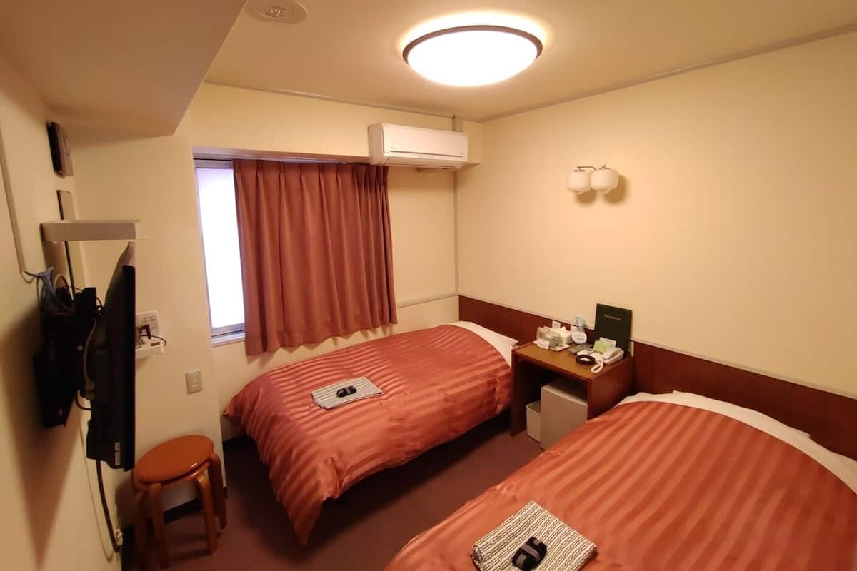Une chambre d’hôtel avec deux lits, un bureau, une chaise, une fenêtre avec des rideaux, un climatiseur et une lampe fixée au mur.