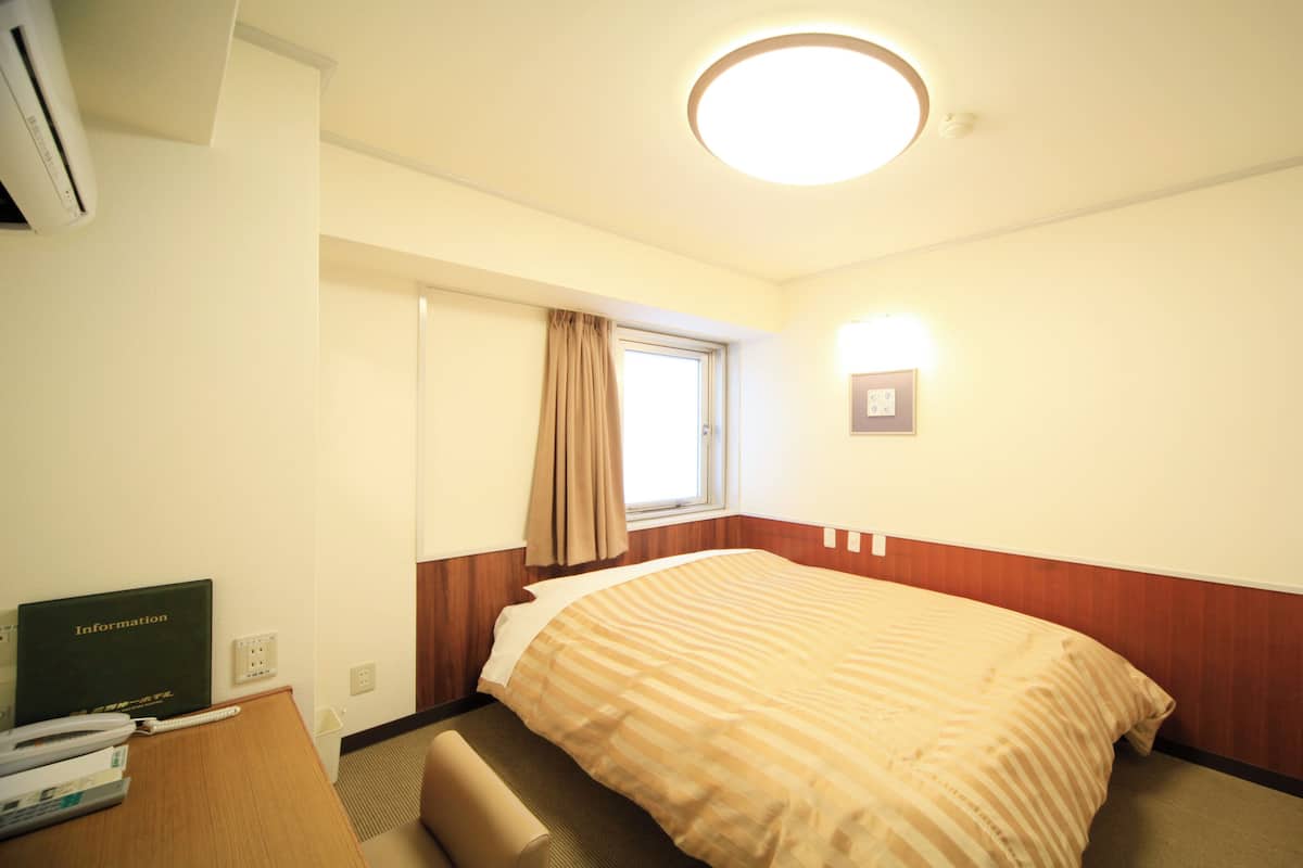 Habitación de hotel con cama, escritorio con teléfono, televisión de pantalla plana, ventana con cortinas e iluminación en el techo.