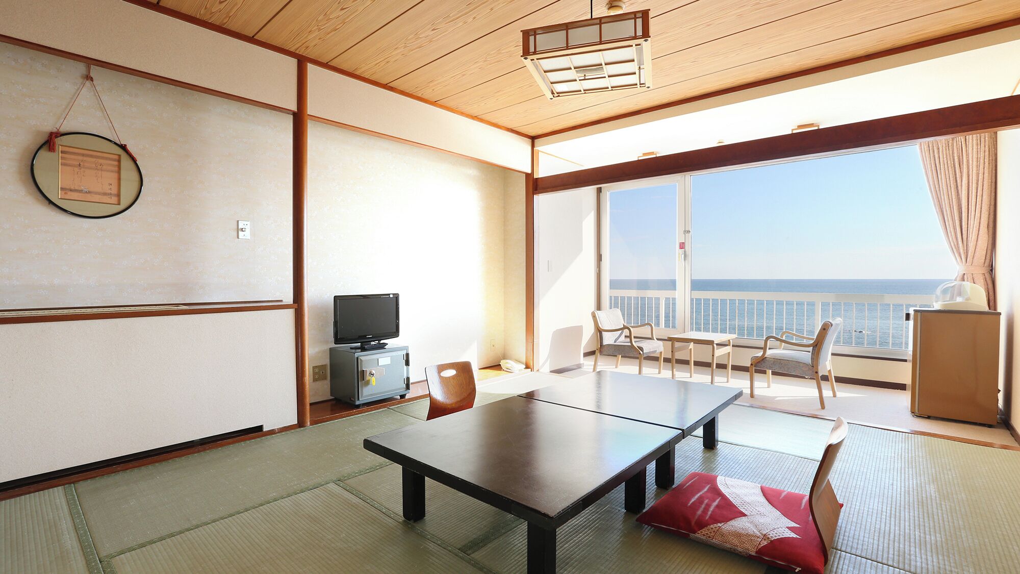 A room with tatami flooring, a television, a table, chairs, and a view of the sea.