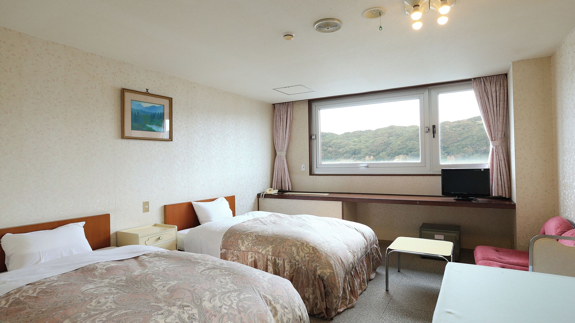 A hotel room with two beds, a desk, a chair, a TV, and a window with a view of hills.