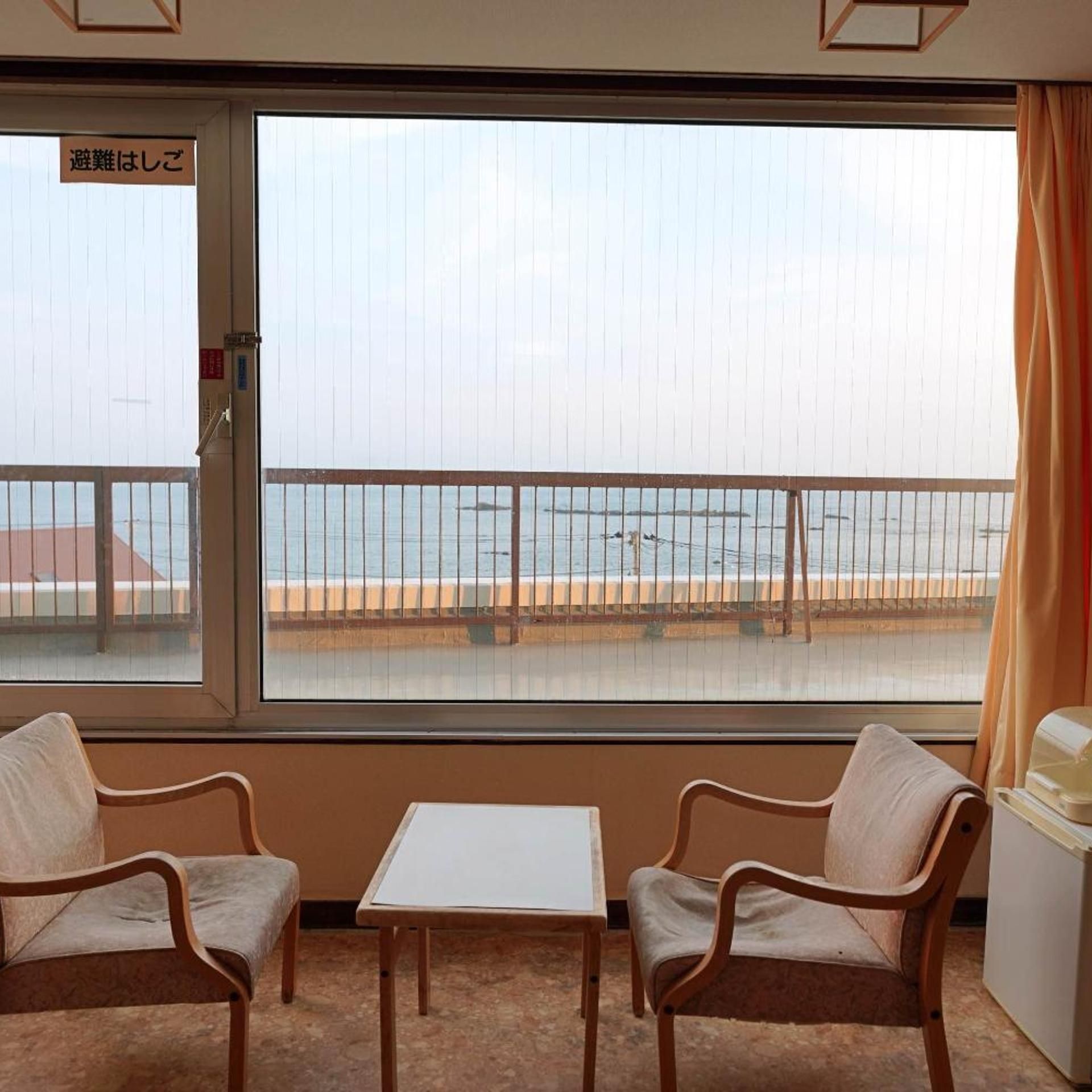 A room with a view of the sea, two chairs, a small table, and a refrigerator.